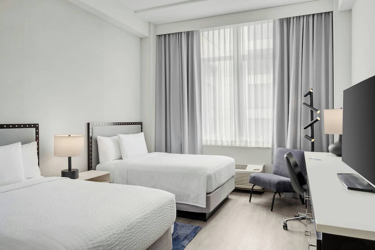 A hotel room with two beds, a desk, a chair, a TV, and a window with curtains.