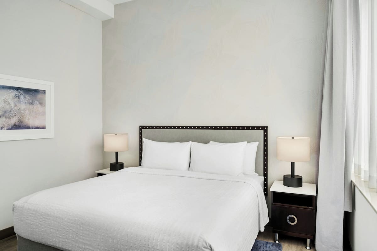 A neatly made bed with white linens, two bedside tables with lamps, a framed picture on the wall, and a window with curtains.