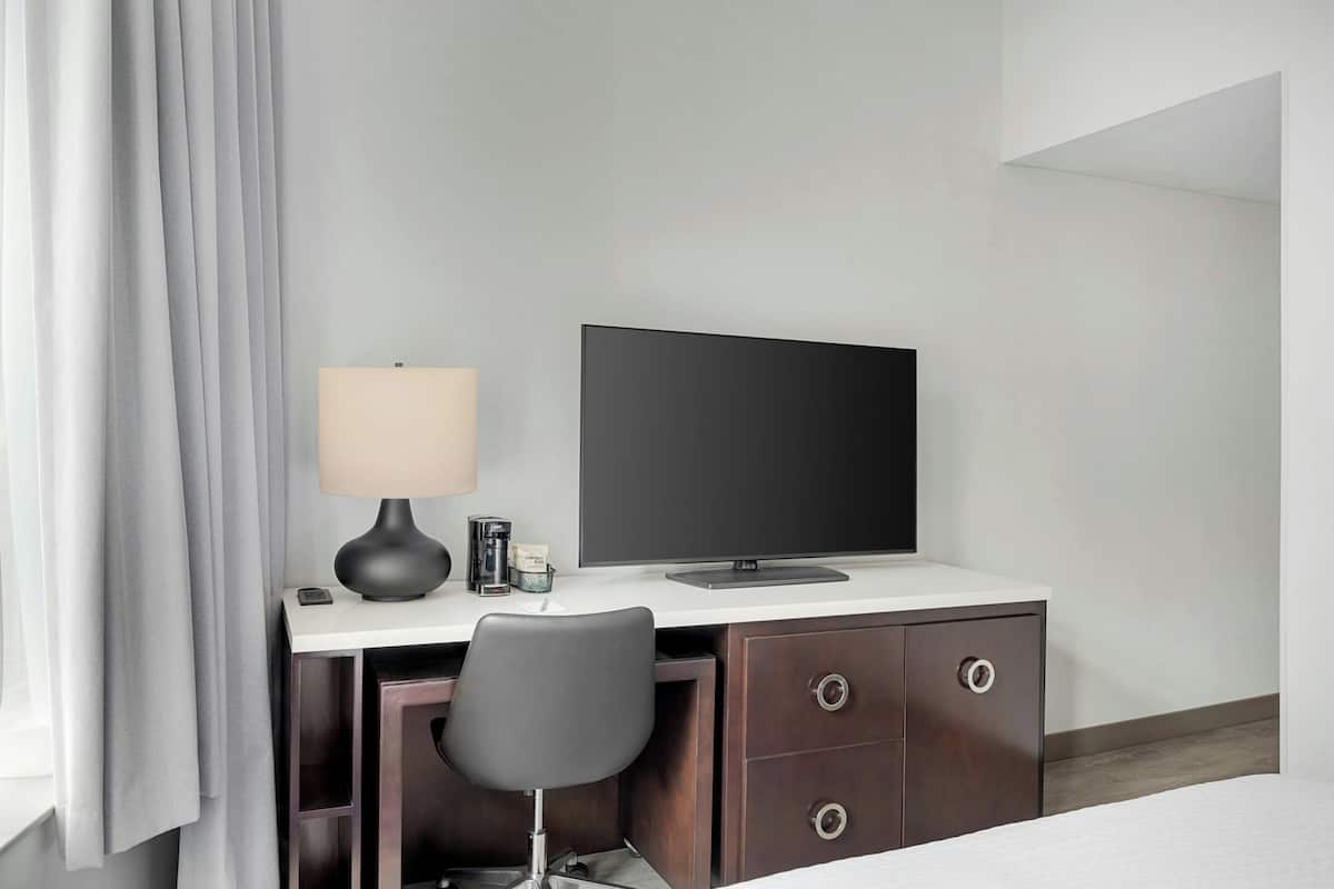 A hotel room with a desk, chair, TV, lamp, and a bed.