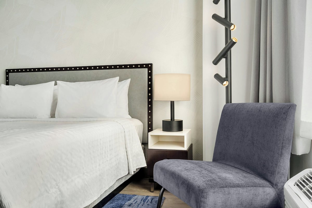 A neatly arranged hotel room with a bed, bedside table with a lamp, a chair, and a wall-mounted light fixture.