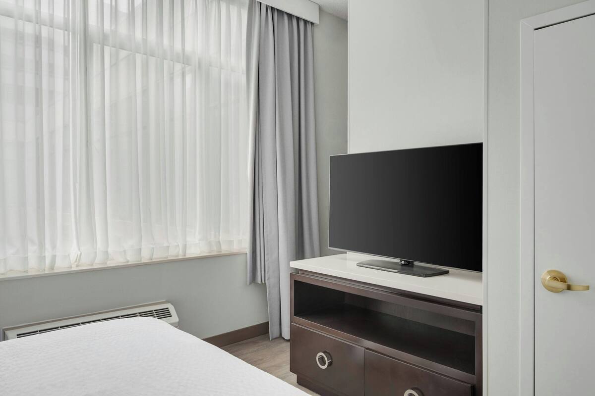 A hotel room with a bed, a television on a white stand, a door with a golden handle, and a window with sheer curtains.