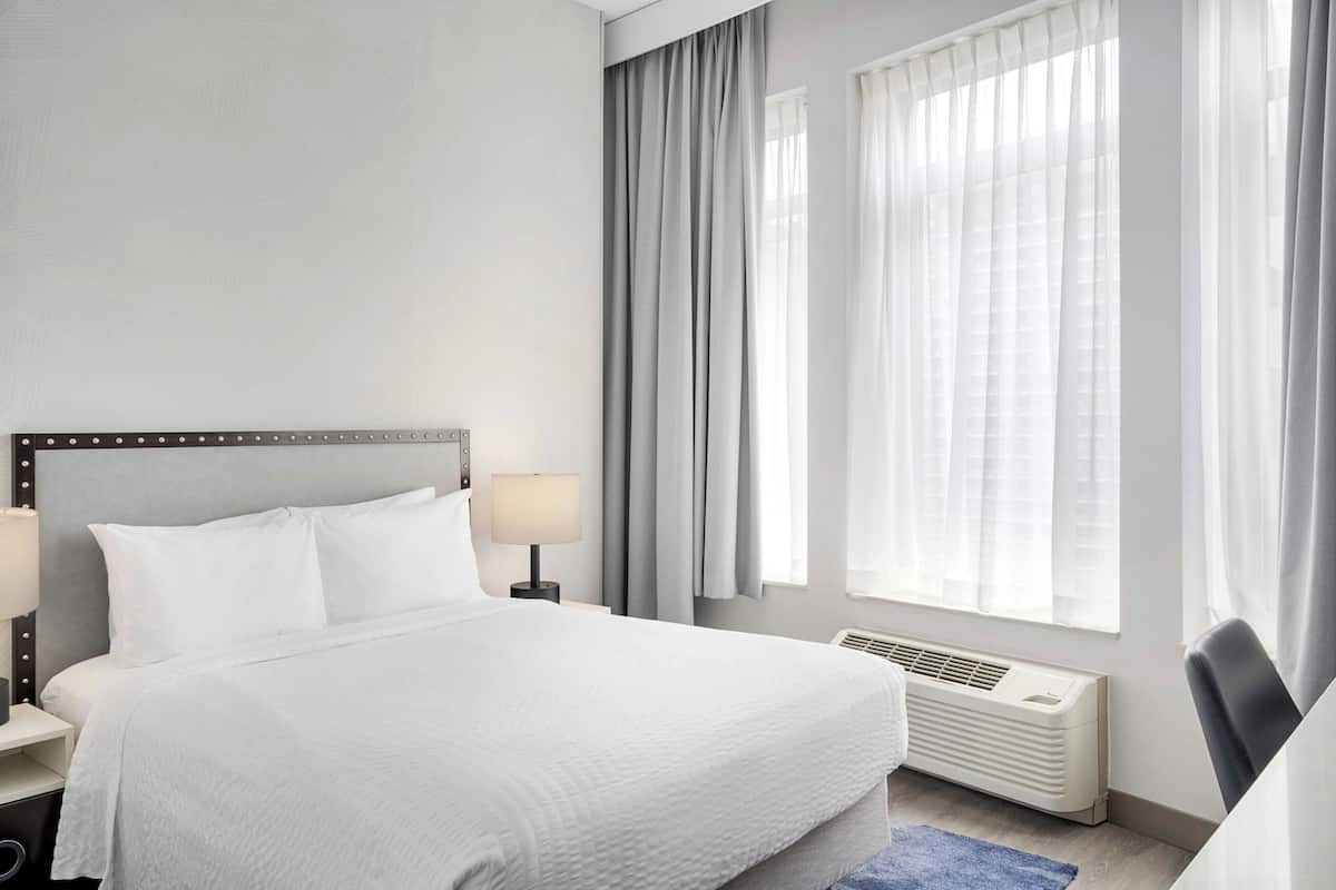 A hotel room with a bed, bedside lamps, a window with curtains, and a radiator.
