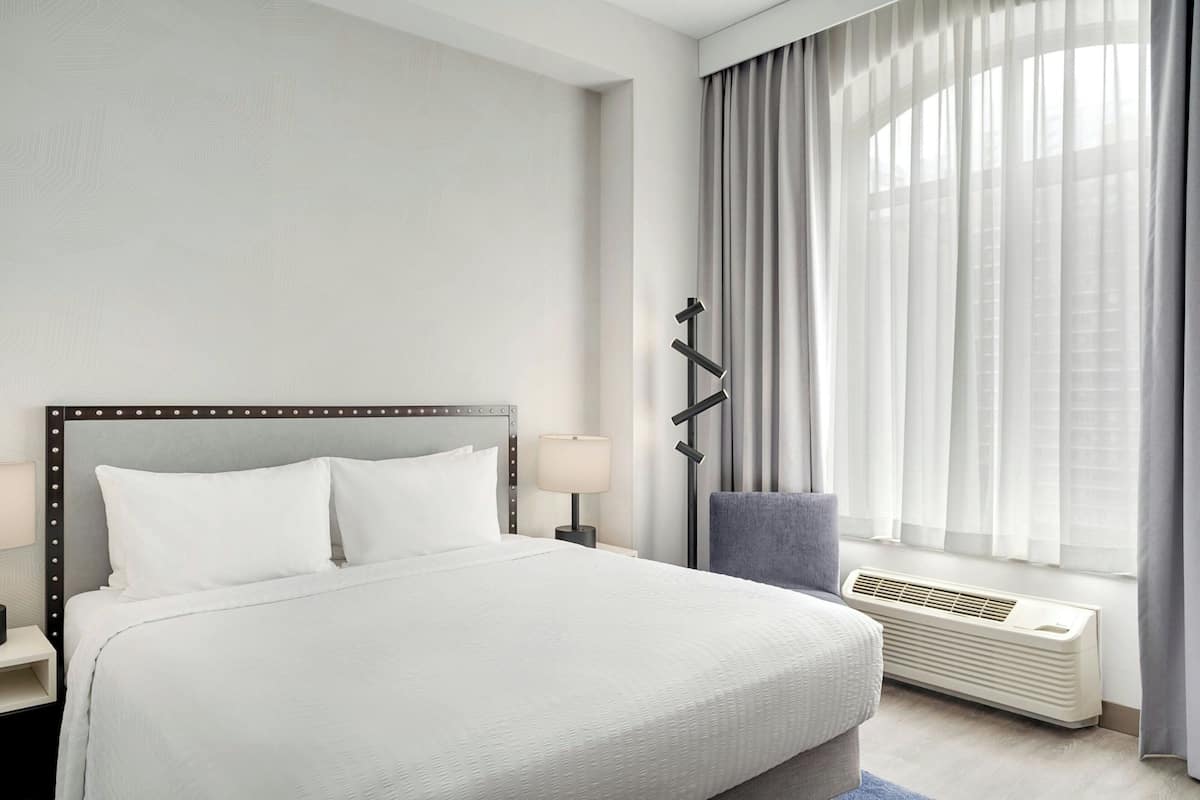 A hotel room with a large bed, a bedside table with a lamp, a chair, a window with curtains, and a wall-mounted air conditioning unit.