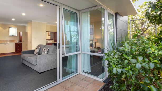 A modern living area with a grey sofa, a glass sliding door, and a view of a kitchen and dining area. - Aristotles North Shore (Auckland)