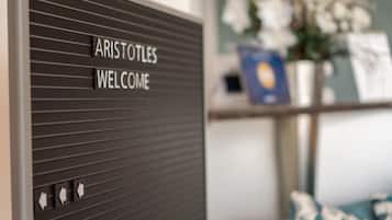 Un letrero con la inscripción "ARISTOTLES WELCOME" en una pared, una estantería con un libro y un jarrón de flores al fondo.