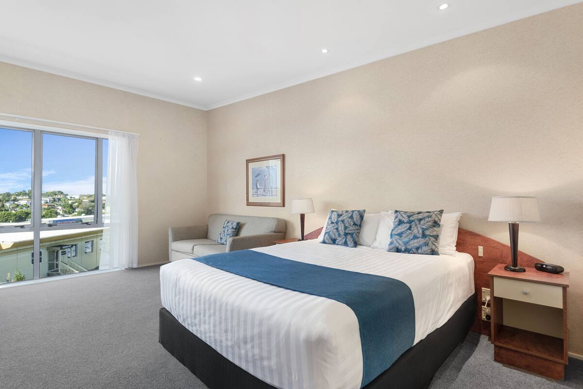 A hotel room with a large bed, a sofa, a desk, and a window with a city view.
