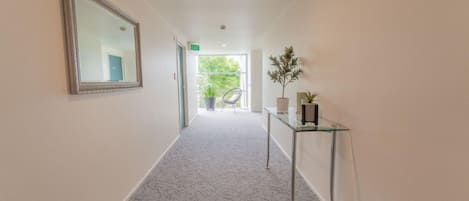 A hallway with a carpeted floor, a mirror on the left, a glass table with a plant on the right, and an exit sign above a door.