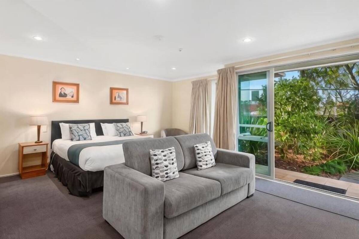A bedroom with a bed, a grey sofa, a bedside table, and a view of greenery through a sliding glass door.