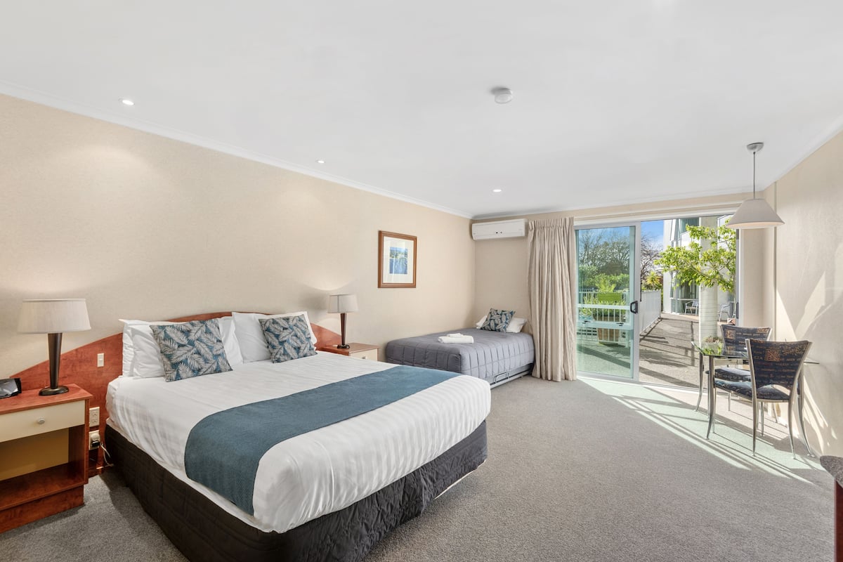 A hotel room with a large bed, a smaller bed, a desk with a lamp, a chair, and a balcony with a view of greenery.