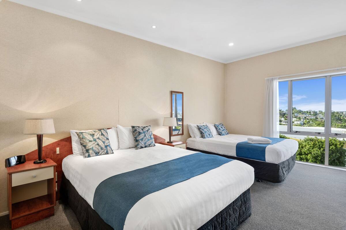 A hotel room with two beds, a nightstand with a lamp, a small table, and a large window offering a view.