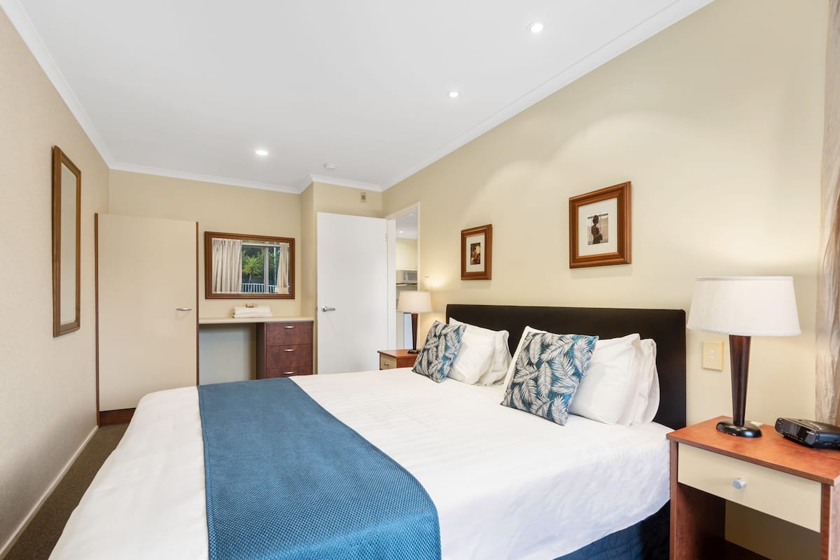 A hotel room with a large bed, a bedside table with a lamp, a small desk with a chair, a mirror, and a picture on the wall.