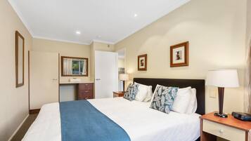 A hotel room with a large bed, a bedside table with a lamp, a small desk with a chair, a mirror, and a picture on the wall.