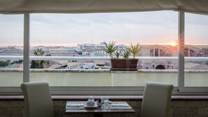 Un coin repas avec une table dressée pour deux personnes, une plante en pot et une vue sur le paysage urbain au coucher du soleil.