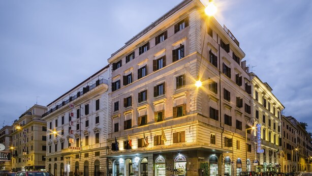 Un hôtel à plusieurs étages, de style architectural classique, doté de balcons et d’une entrée au niveau du sol, avec des boutiques.