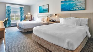 Habitación de hotel con dos camas, un escritorio, una silla, un ventanal con cortinas y obras de arte en las paredes.