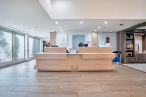 A reception desk with a modern design, a blue door in the background, and a view of the outside through large windows. - Hilton Garden Inn Jacksonville Airport (Jacksonville)