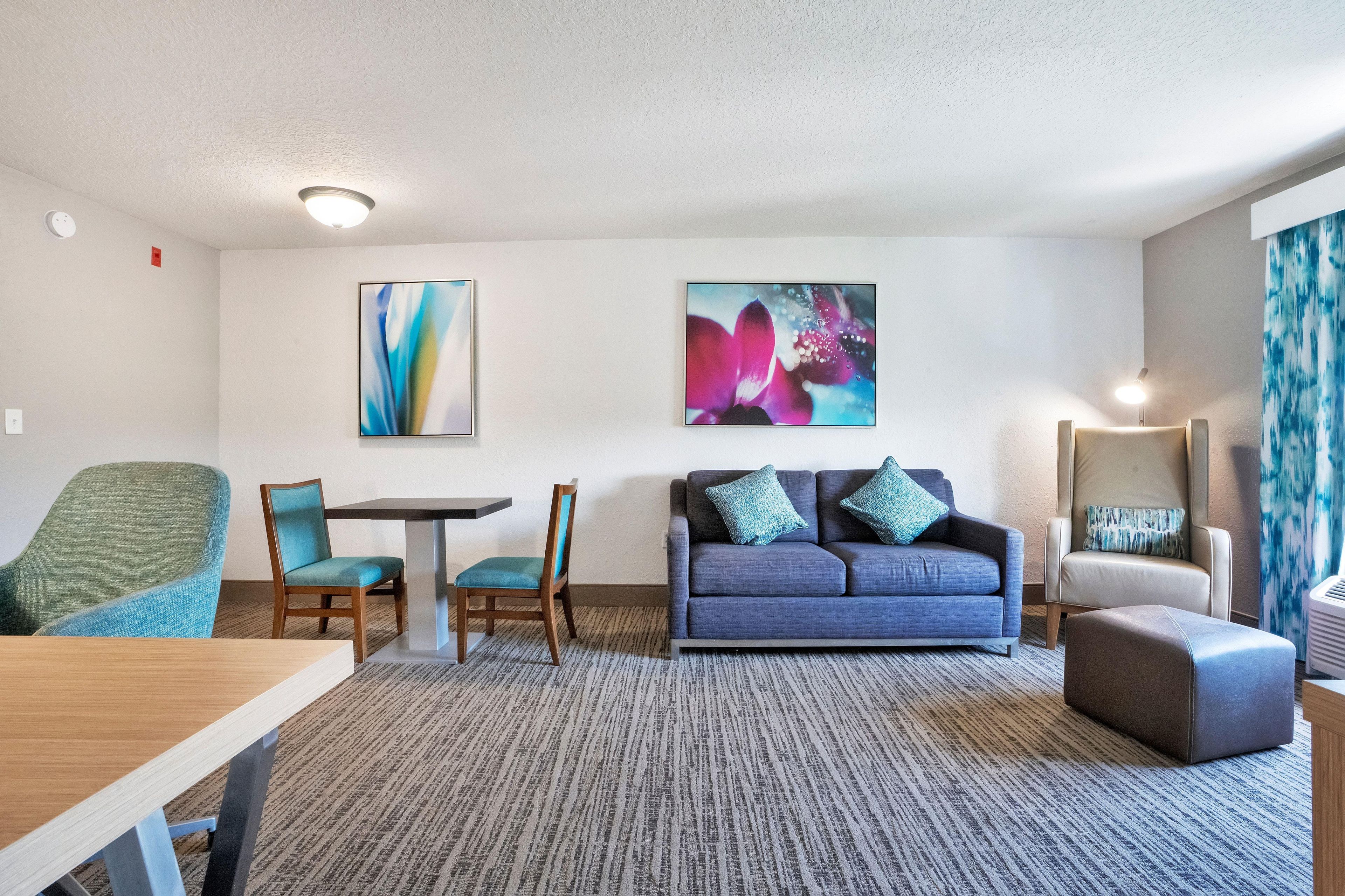 A hotel room with a dining table, two chairs, a sofa with pillows, a standing lamp, and abstract wall art.