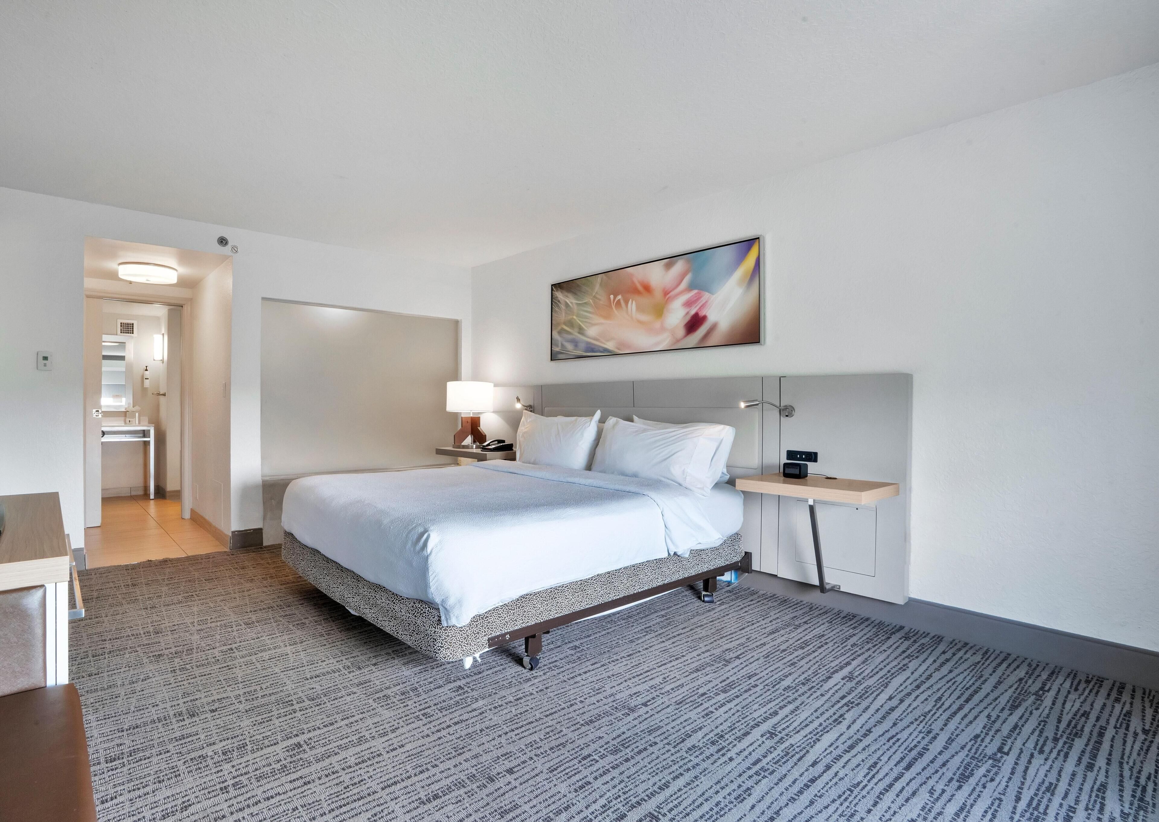 A hotel room with a bed, bedside table, lamp, and a painting on the wall.