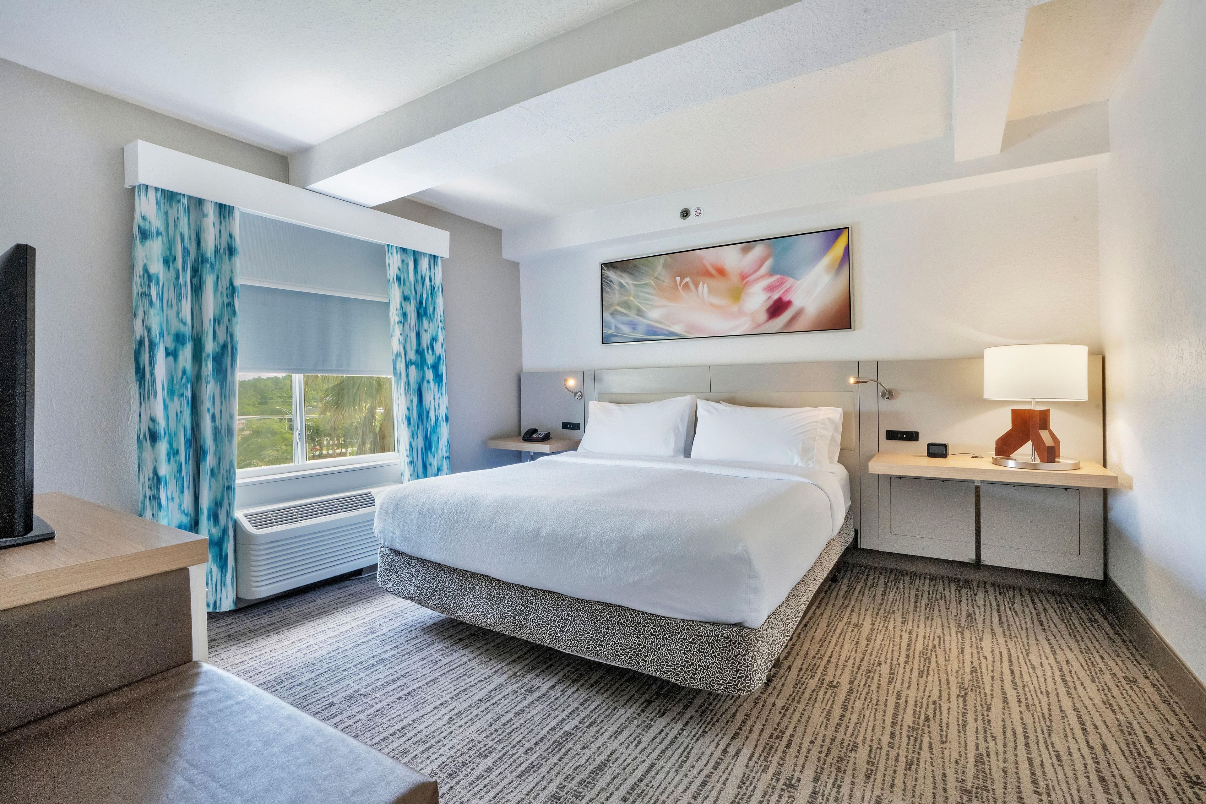 Habitación de hotel con una cama grande, un escritorio, una silla, un televisor, una ventana con cortinas, un cuadro en la pared y una lámpara.