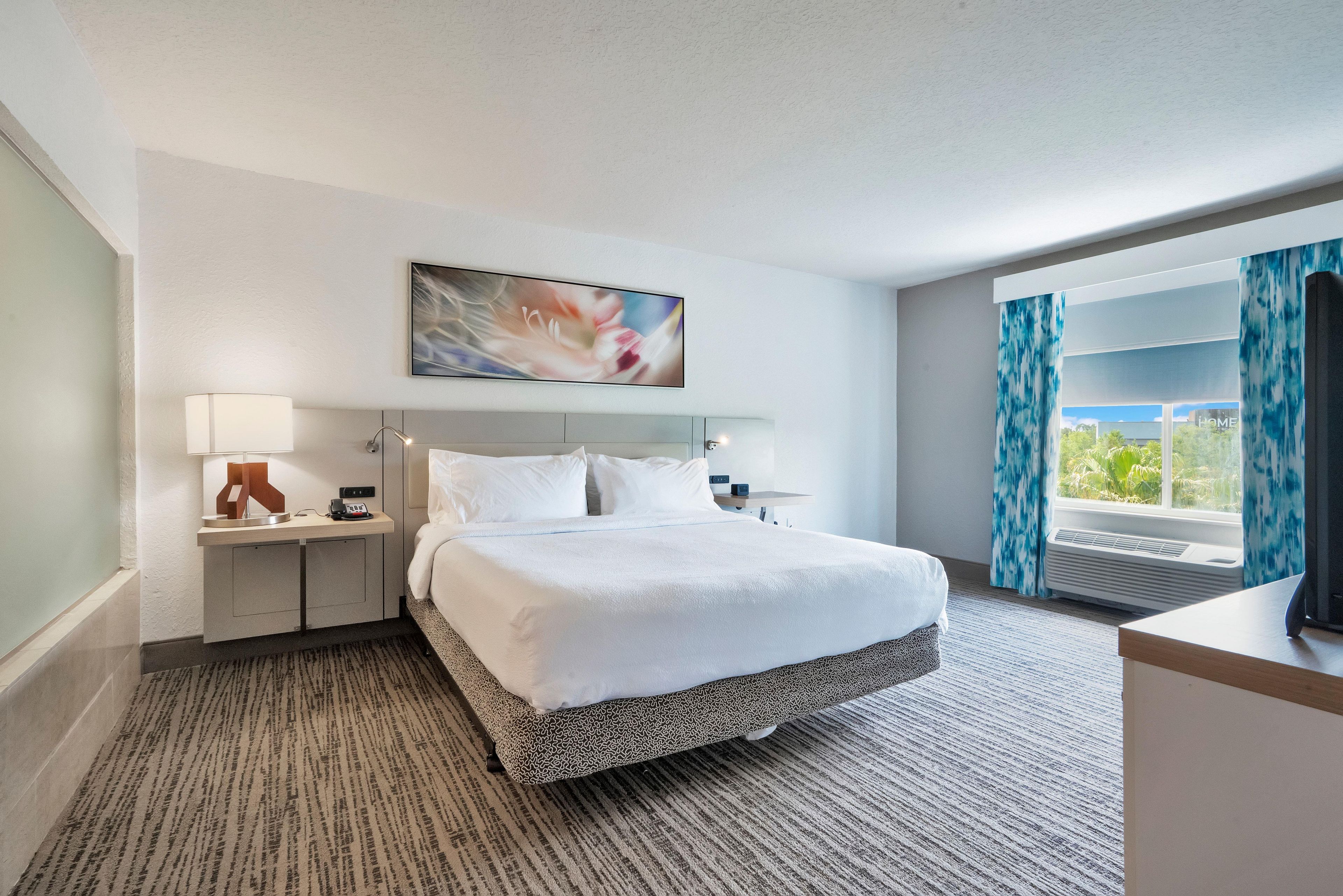 Une chambre d’hôtel avec un lit, une table de chevet, une lampe, un tableau, une fenêtre avec des rideaux et une vue sur la verdure à l’extérieur.