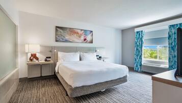 Une chambre d’hôtel avec un lit, une table de chevet, une lampe, un tableau, une fenêtre avec des rideaux et une vue sur la verdure à l’extérieur.
