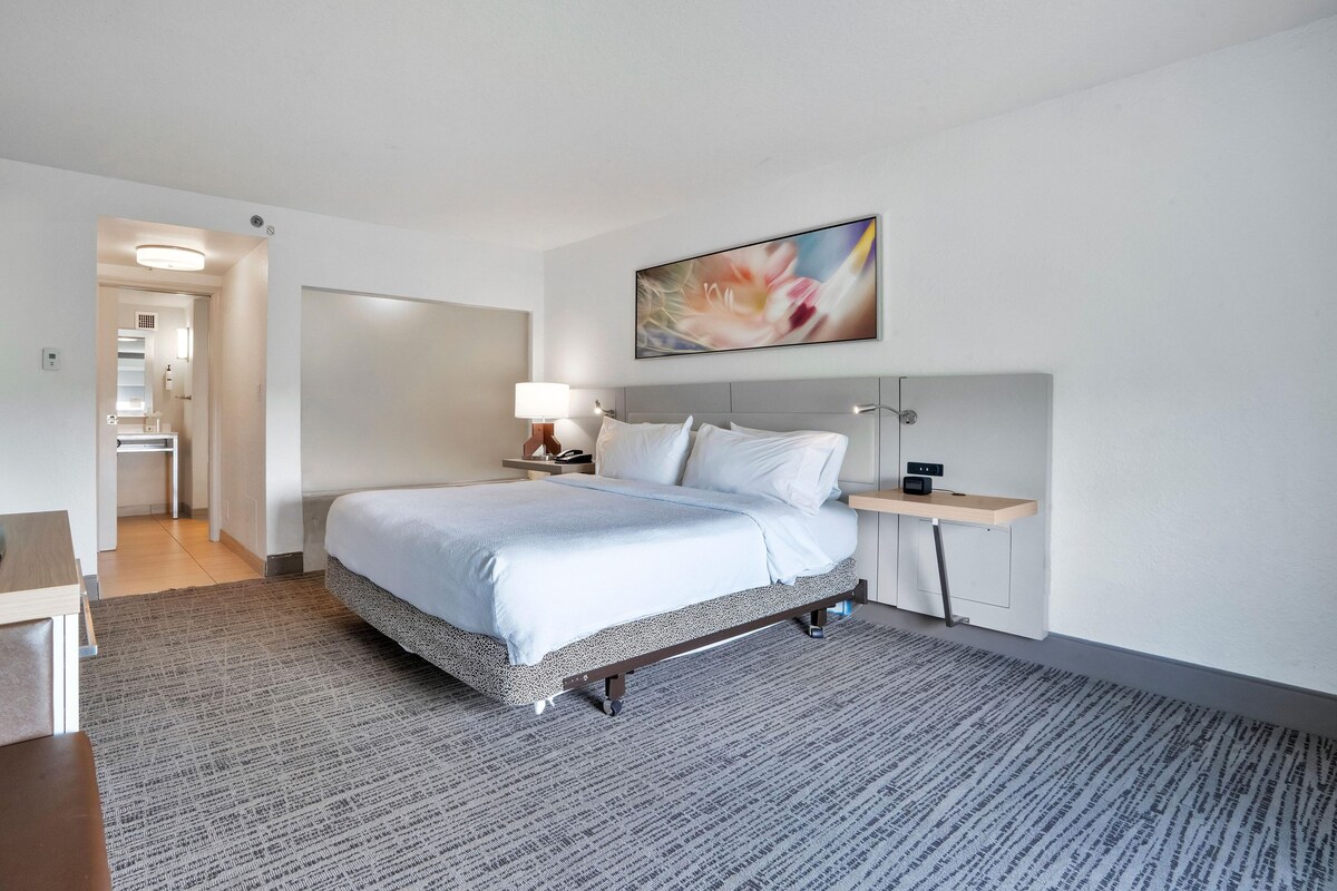 A hotel room with a bed, bedside table, lamp, and a painting on the wall.