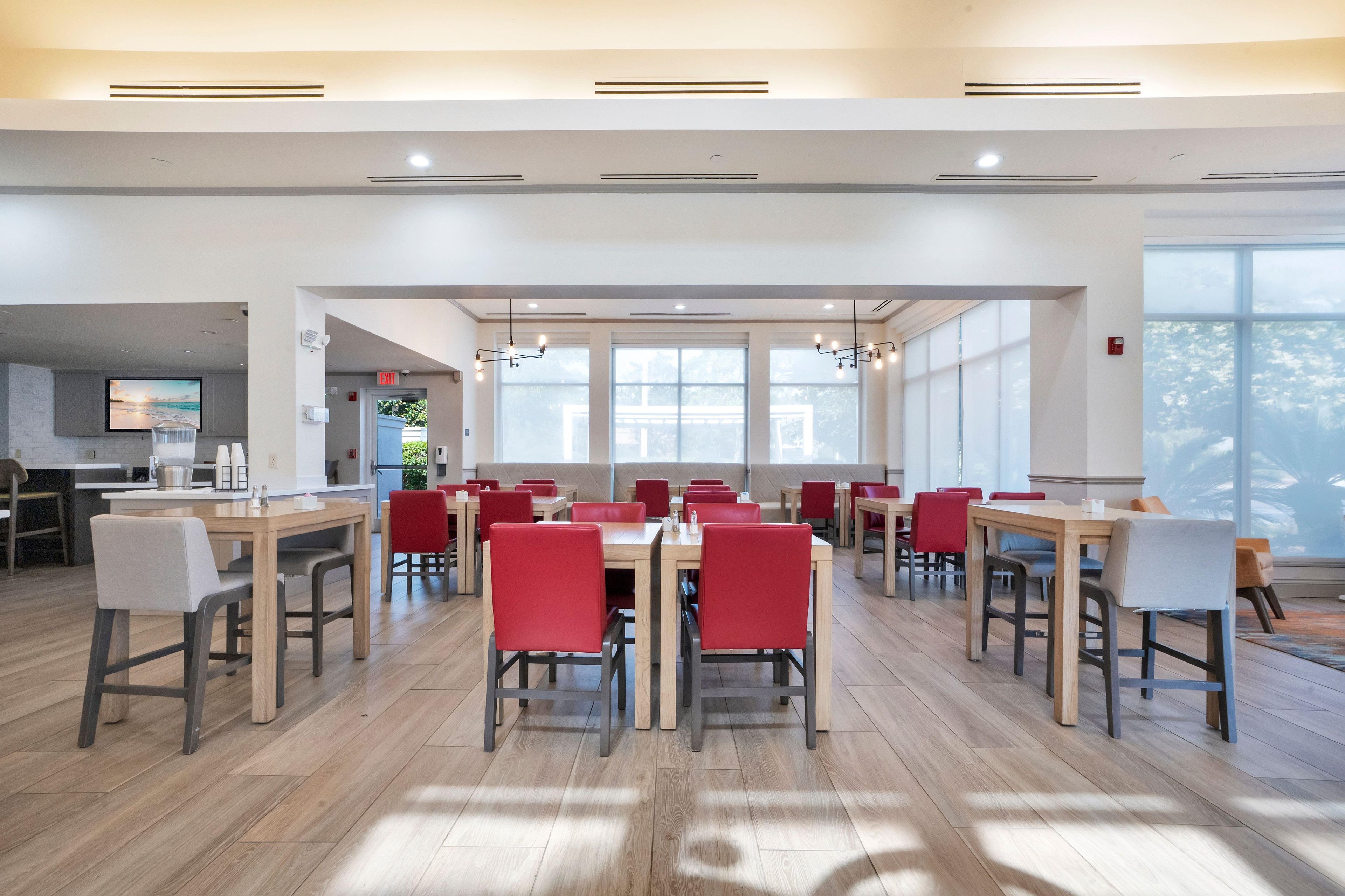 A spacious dining area with multiple tables and chairs, large windows allowing natural light, and a visible kitchen area in the background.