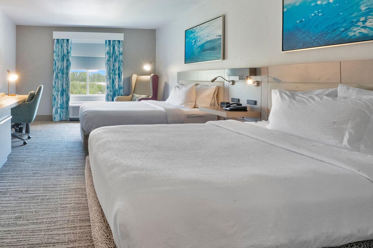 A hotel room with two beds, a desk, a chair, a window with curtains, a painting, and a nightstand with a lamp.