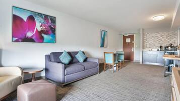 Een moderne woonkamer met een grijze bank, teal kussens, een klein rond tafeltje en een beige fauteuil. Achtergrond toont een keuken met een magnetron en een koffiezetapparaat.