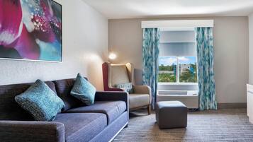 Een moderne hotelkamer met een grijze bank, twee blauwe kussens, een klein voetenbankje, een vloerlamp en een groot raam met gordijnen.