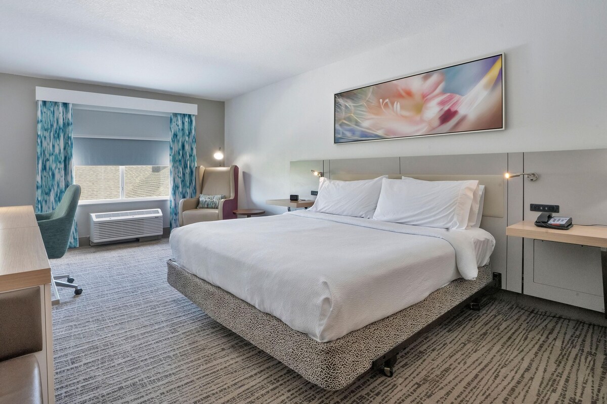 A hotel room with a large bed, a desk, a chair, a window with curtains, and a painting on the wall.