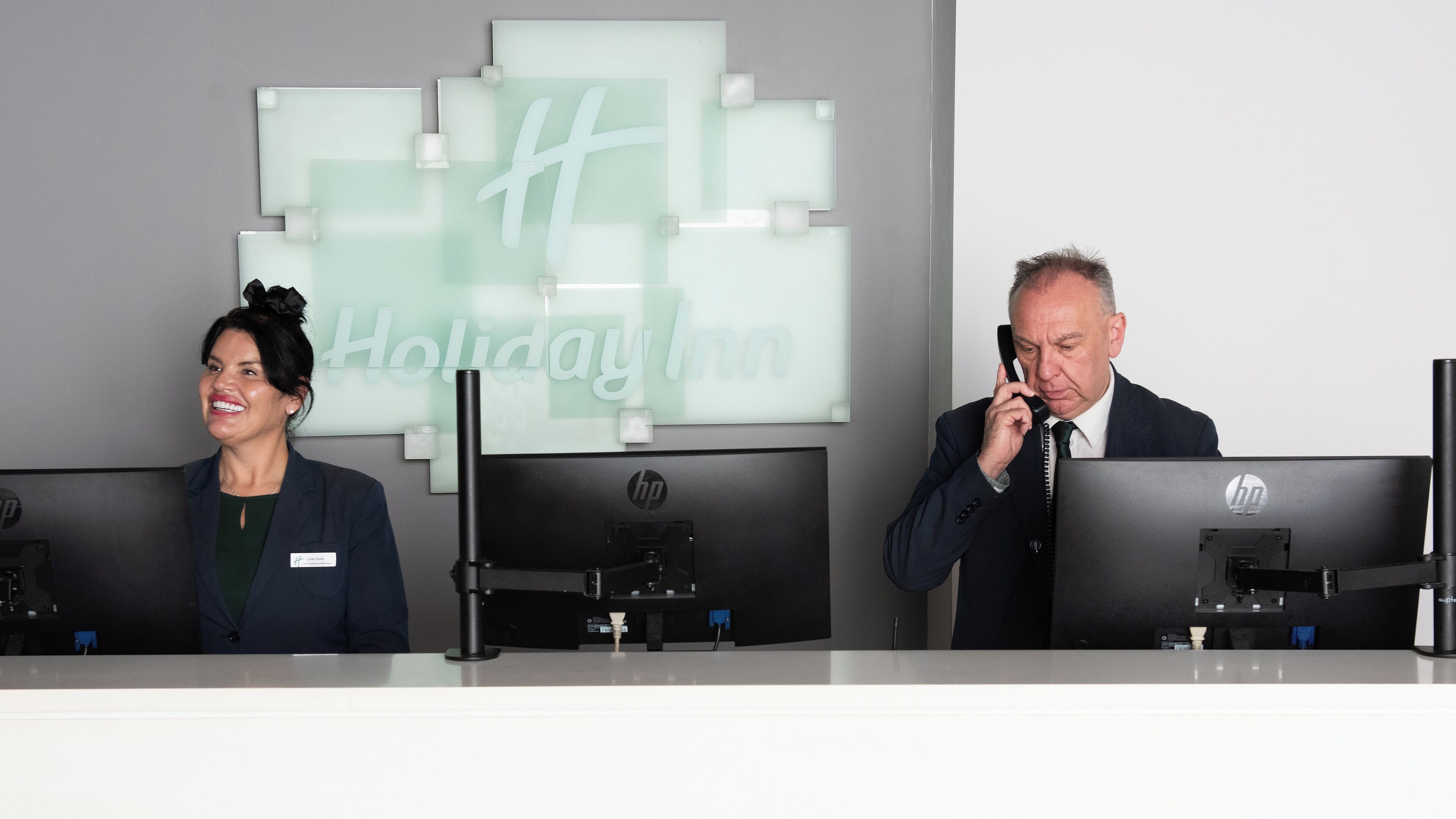 Duas pessoas no balcão de recepção com computadores HP, uma placa da Holiday Inn e um design de interiores moderno.