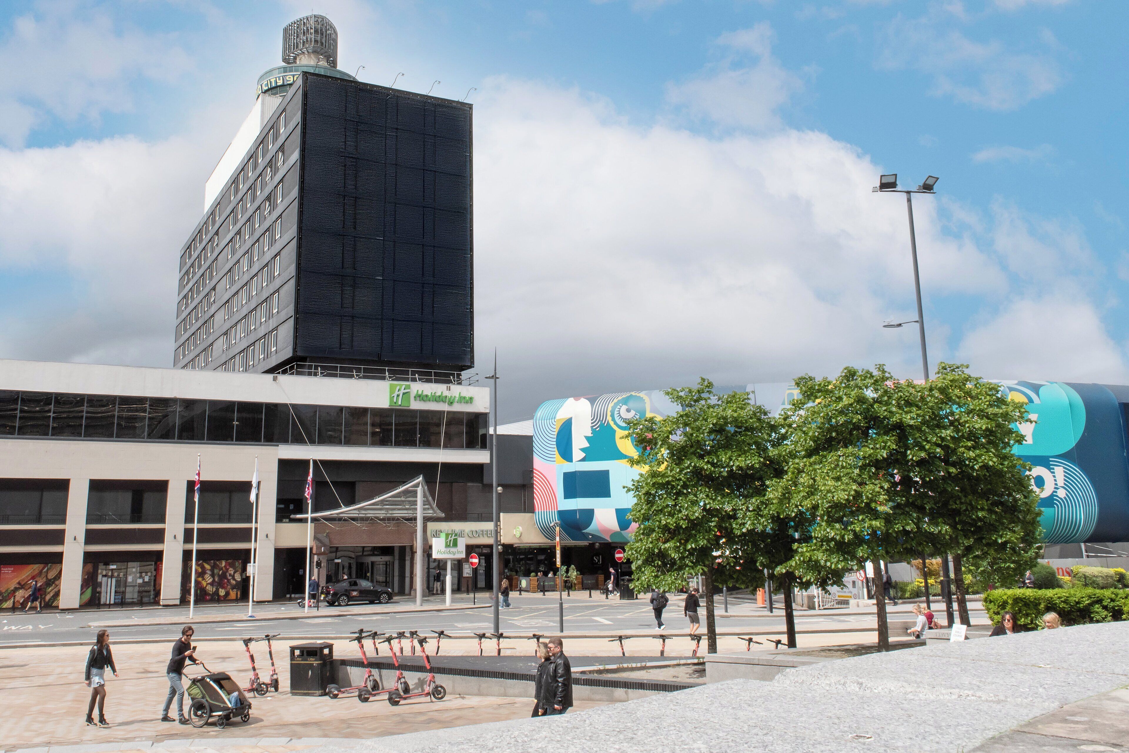 A modern building with a large glass facade, a prominent entrance, and a surrounding plaza with trees and benches.