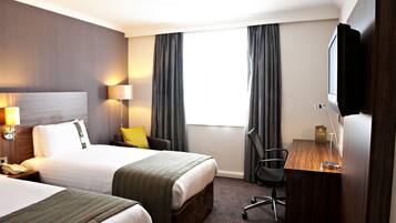 A hotel room with two beds, a desk, a chair, a TV, and a lamp.