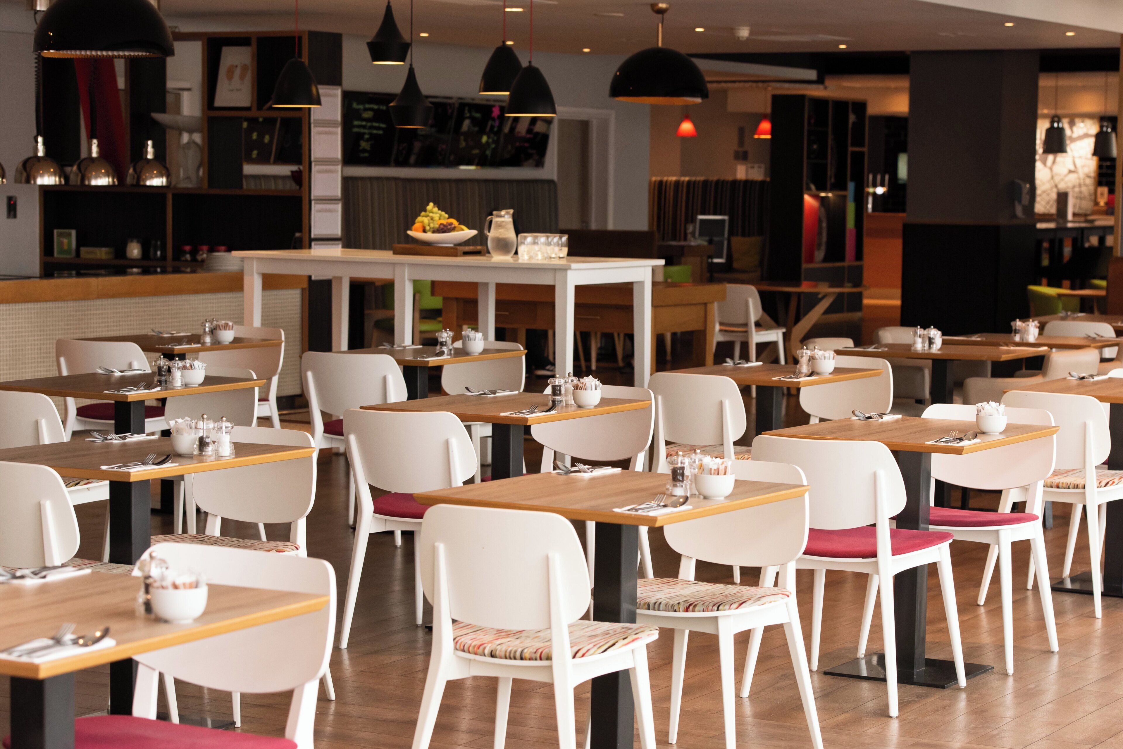 Un restaurant moderne avec des chaises blanches et des tables en bois, agrémenté de luminaires suspendus et doté d’un espace central pour le service.