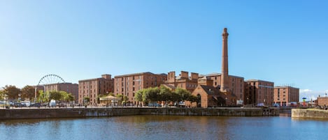 A waterfront hotel with a prominent chimney, a Ferris wheel in the background, and a calm river in the foreground.