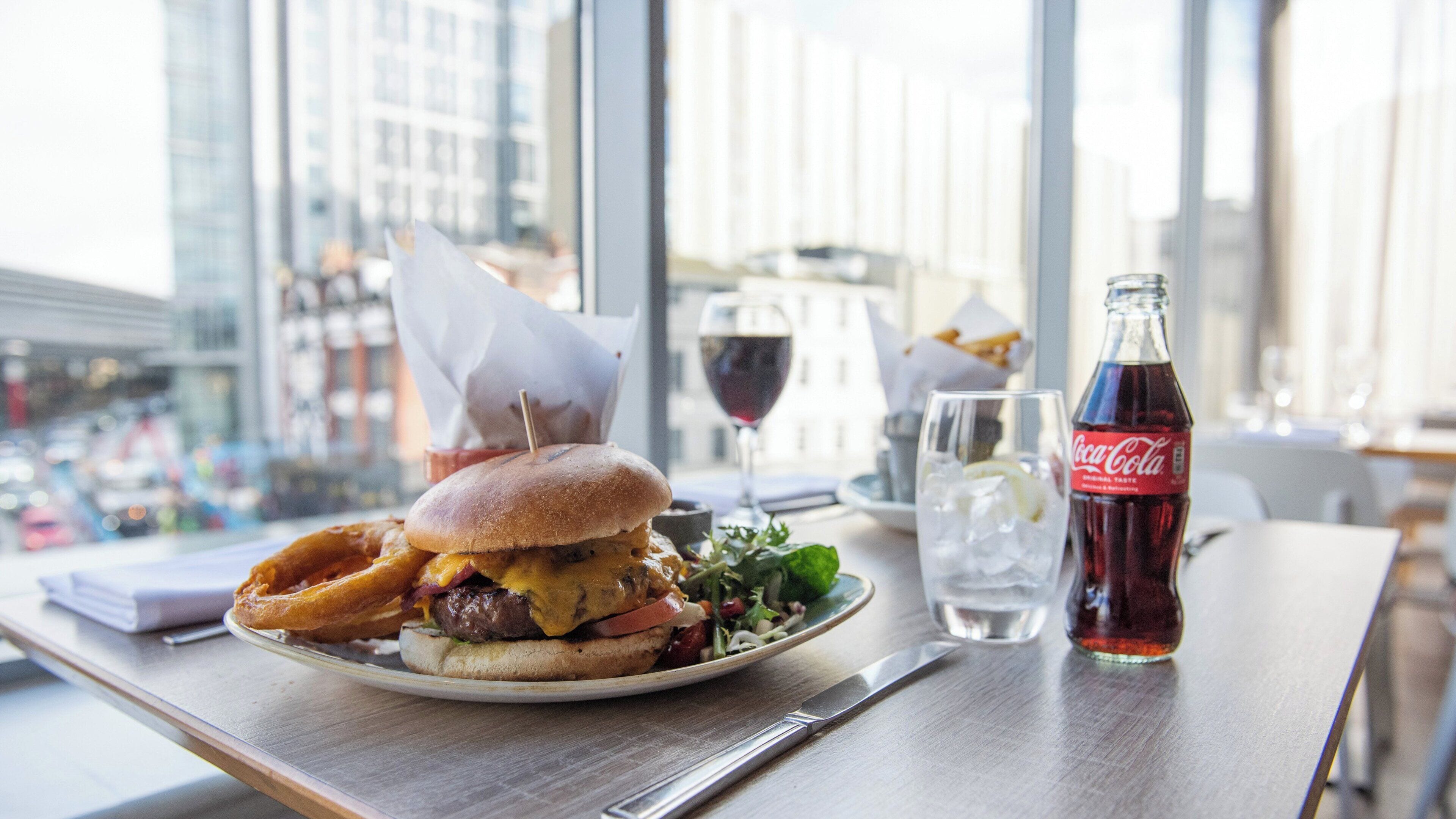 A meal with a burger, fries, and a salad on a plate, a glass of wine, and a bottle of Coca-Cola on a table with a view of buildings through large windows.