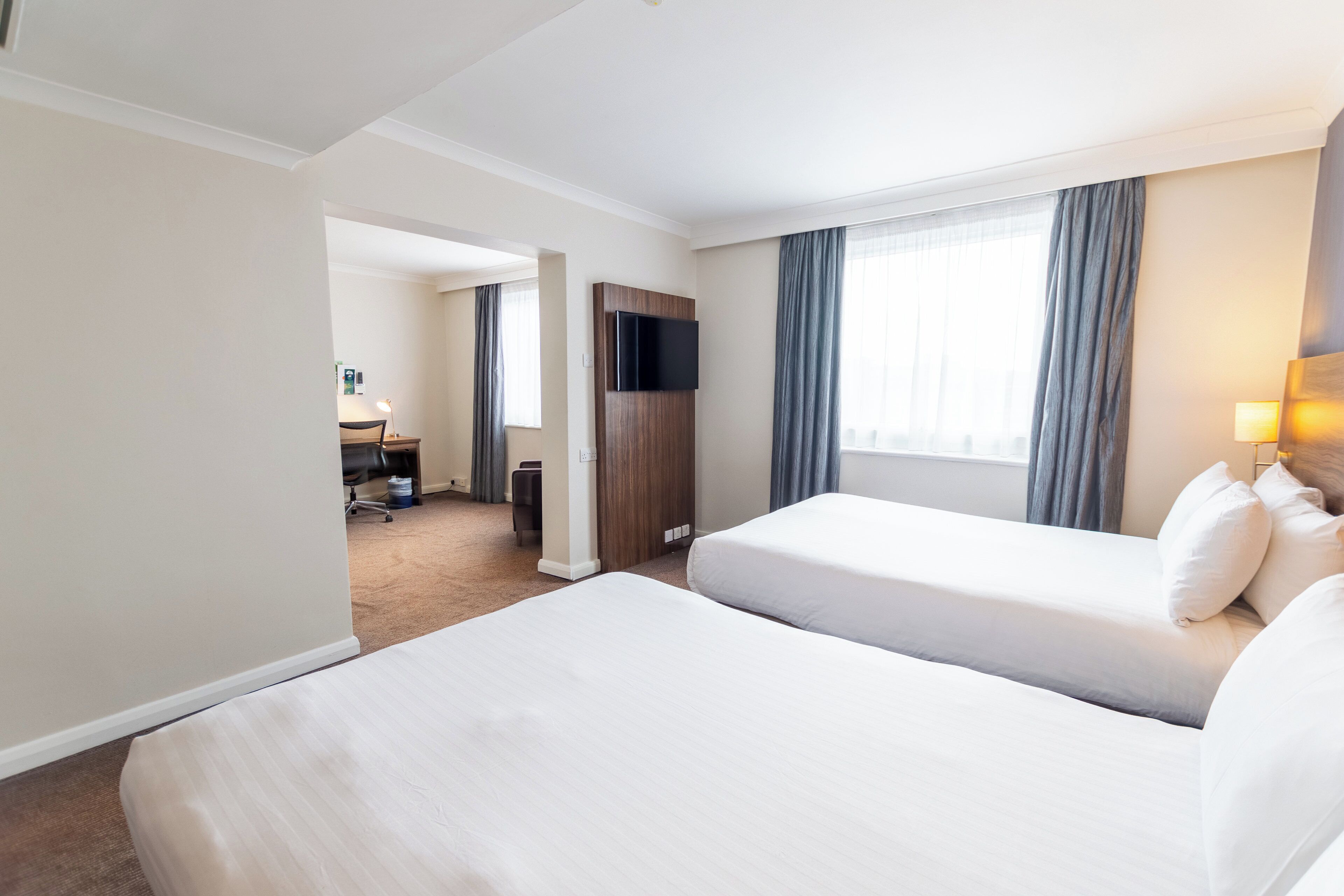 Ein Hotelzimmer mit zwei Betten, einem Schreibtisch, einem Stuhl, einem Fernseher und einem Fenster mit Vorhängen.