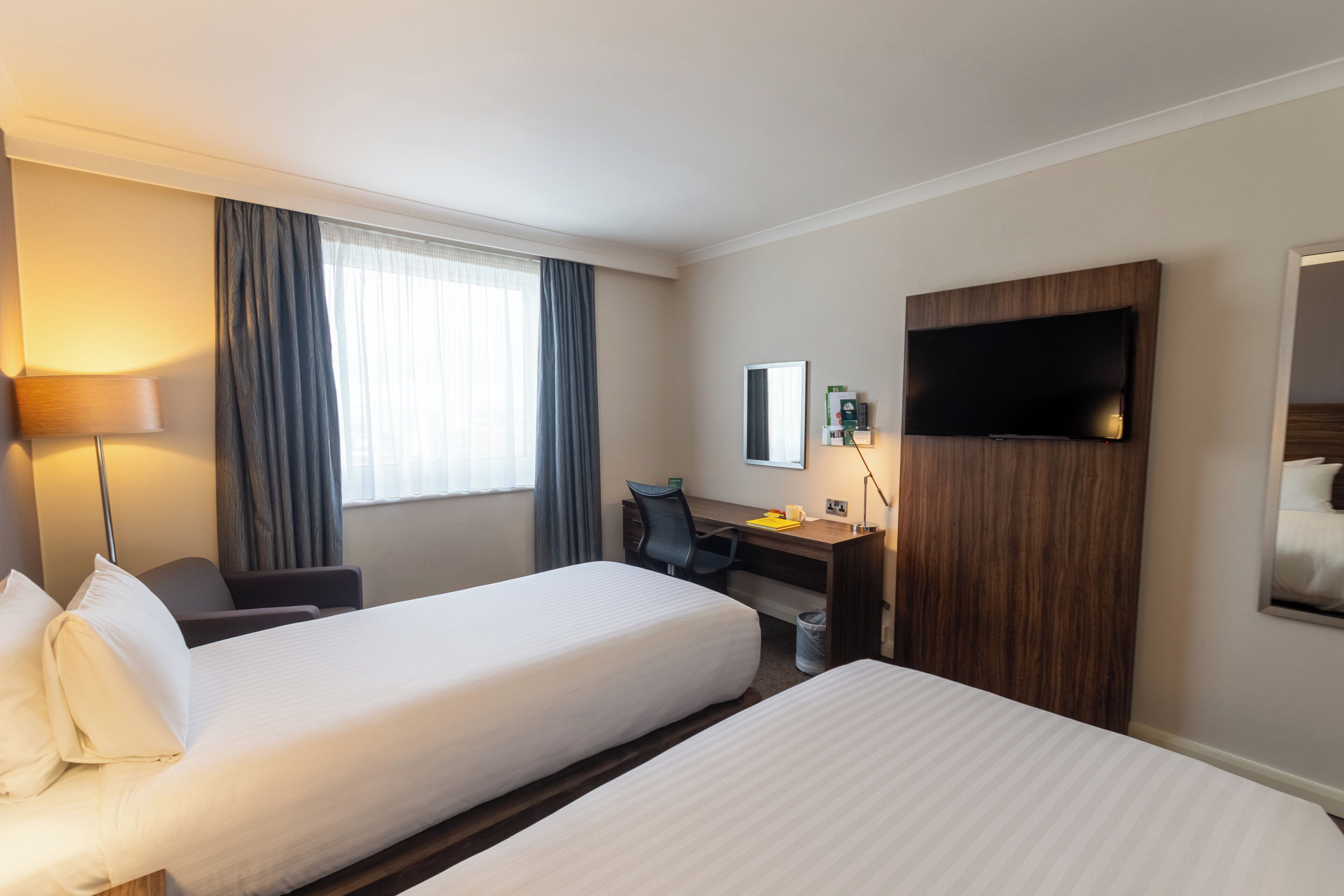 A hotel room with two beds, a desk, a chair, a TV, and a window with curtains.