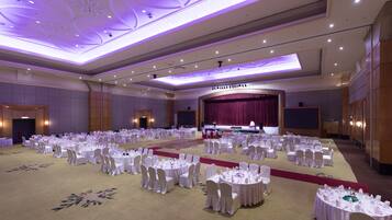 A large banquet hall with round tables and chairs, a stage with a red curtain, and a carpeted floor.