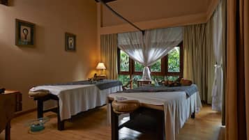 A room with two beds, a massage table, a window with curtains, and a small table with a lamp.