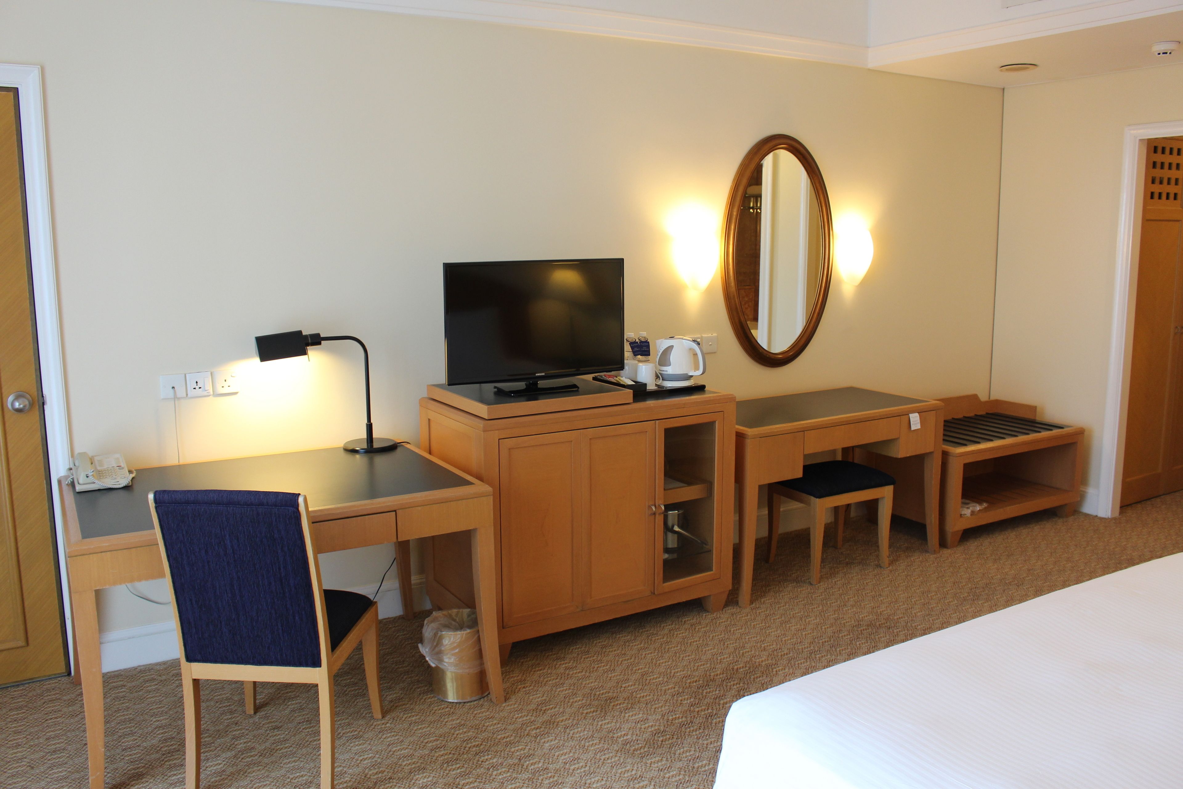 A hotel room with a bed, a desk with a computer, a chair, a television, a mirror, and a lamp.
