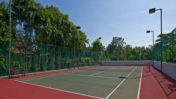 Una cancha de tenis exterior con superficie roja, rodeada por una cerca verde y palmeras.
