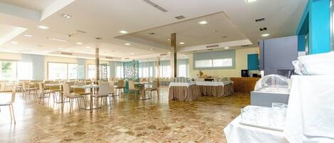 Une salle de conférence spacieuse dotée d’un sol en pierre polie, de plusieurs tables recouvertes de nappes blanches et d’un espace buffet avec de la vaisselle en verre transparent.
