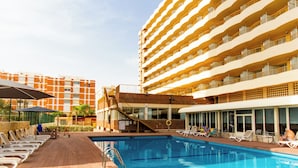 Zona junto a la piscina con sillones de descanso y una pileta, rodeada por un edificio de hotel de varios pisos.