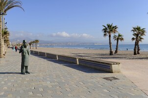 Un sendero pavimentado con bancos y palmeras que conduce a una playa con una estatua y una vista despejada del océano y montañas lejanas.