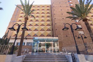 Una entrada de hotel con fachada geométrica, palmeras y acceso de vidrio.