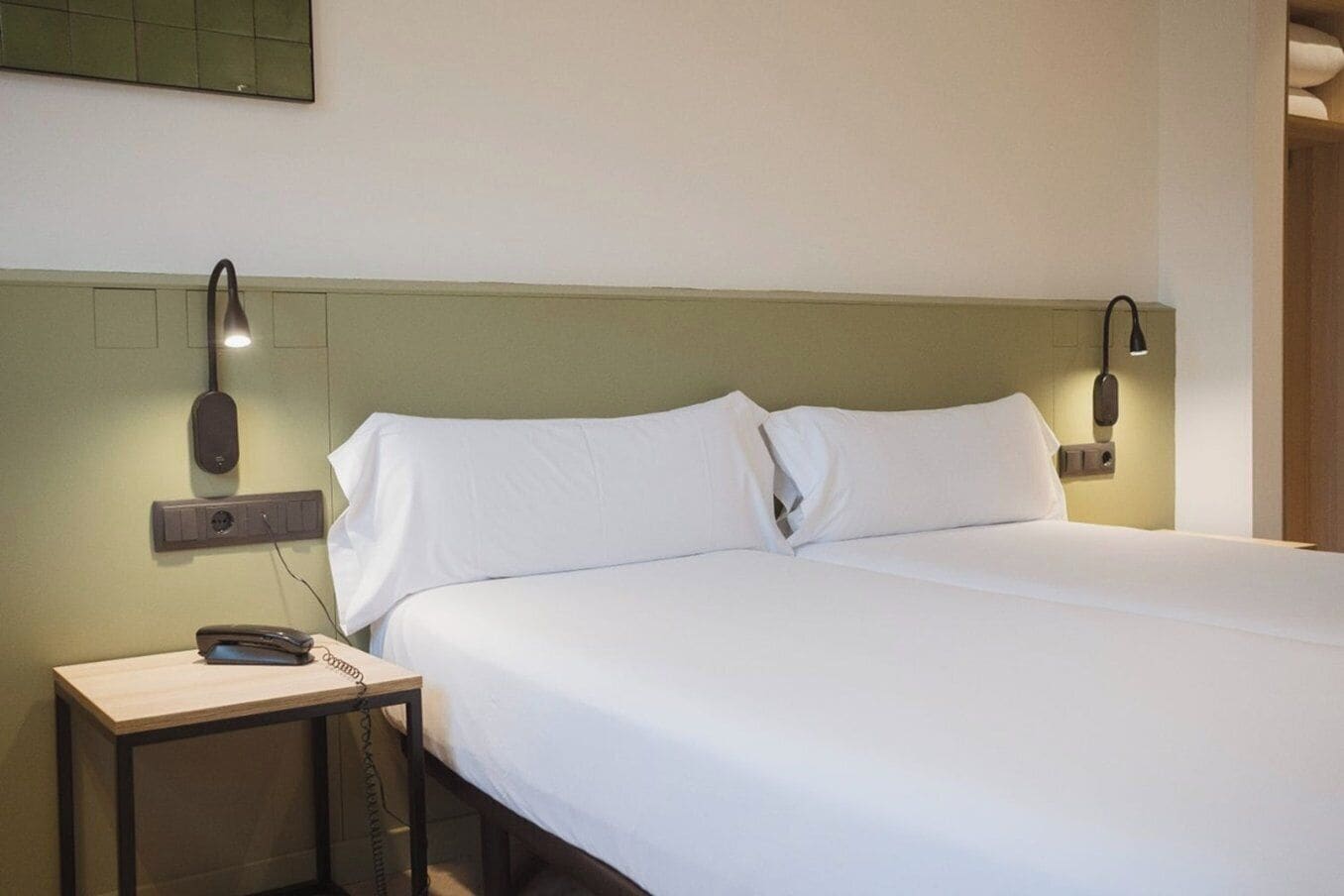 Een netjes opgemaakt bed met witte lakens, een nachtkastje met een lamp en een aan de muur bevestigde telefoon.