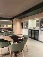 A modern dining area with green chairs and tables, a kitchen counter with a refrigerator, and a wall-mounted TV.