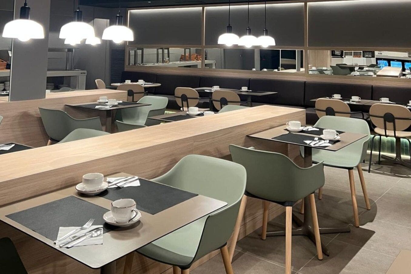 Een modern restaurant met lange gedeelde tafels en individuele zitjes, hangende verlichting en een grote spiegel die het interieur weerkaatst.